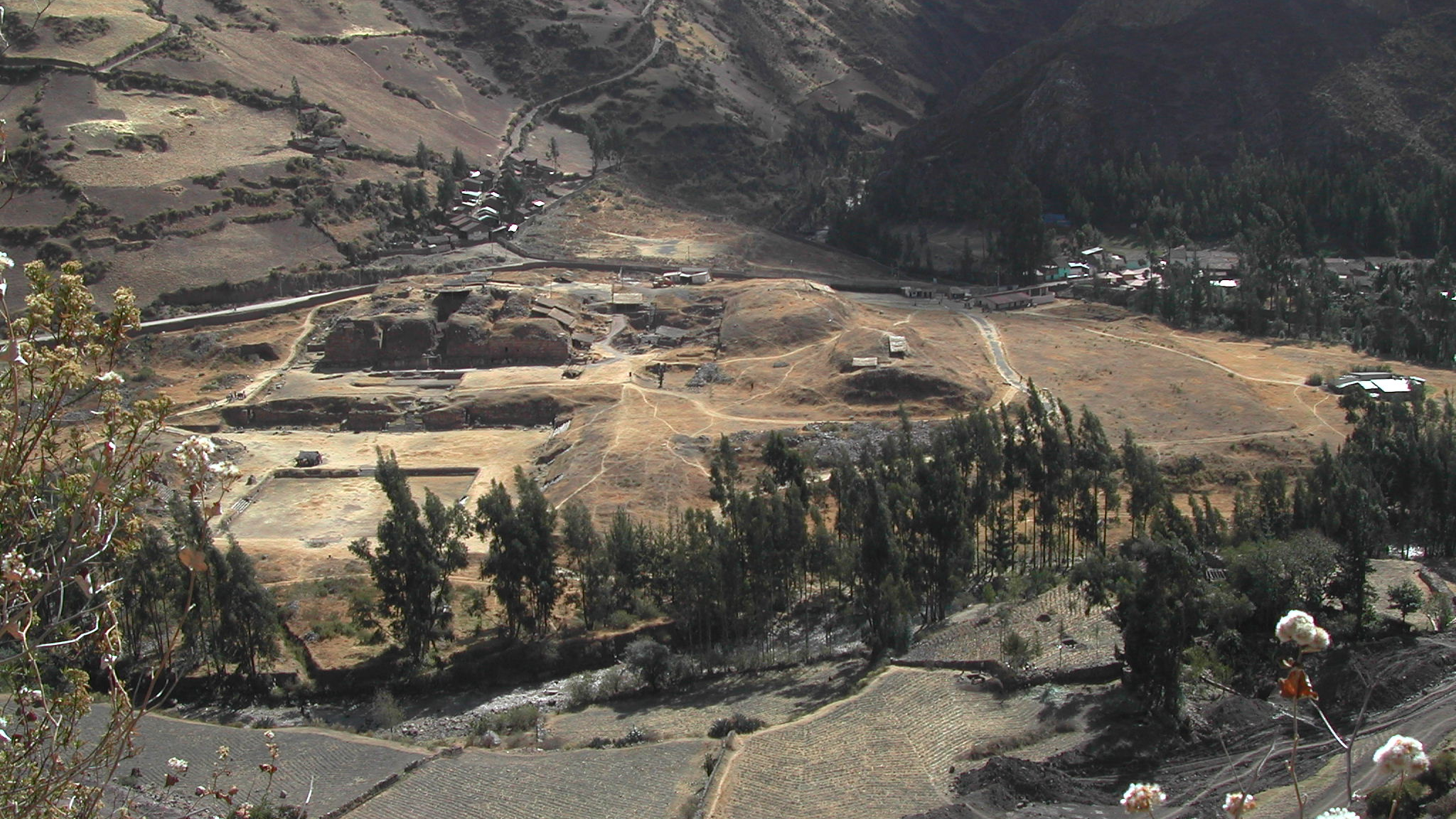 View of Chavín archaeological site in Peru. The ground is brown with scrubby trees and mountains rising in the background.