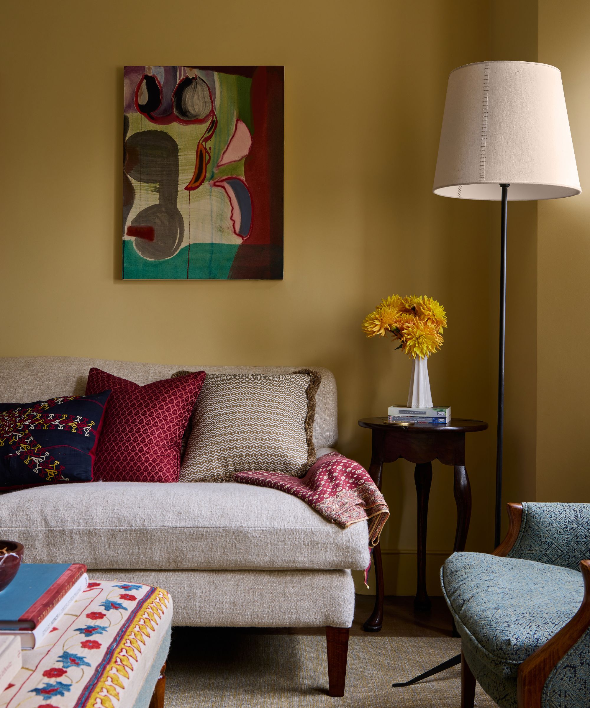 Yellow liivng room with neutral linen sofa, colourful patterned cushions, an abstract maroon and green painting, a side table with a vase of yellow flowers, and a large floor lamp with a cream shade.