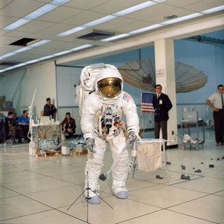 As part of training exercises at NASA's Kennedy Space Center in Florida, astronaut Jim Lovell, commander of the Apollo 13 lunar landing mission, simulates an extravehicular activity on the lunar surface — also known as a "moonwalk" — while inside the Flight Crew Training Building. Lovell is holding an Apollo Lunar Hand Tool in his left hand and is wearing an Extravehicular Mobility Unit (EMU) spacesuit. In front of Lovell is a gnomon (a sundial-like tool on a tripod), and behind him to the right is a tool carrier.
