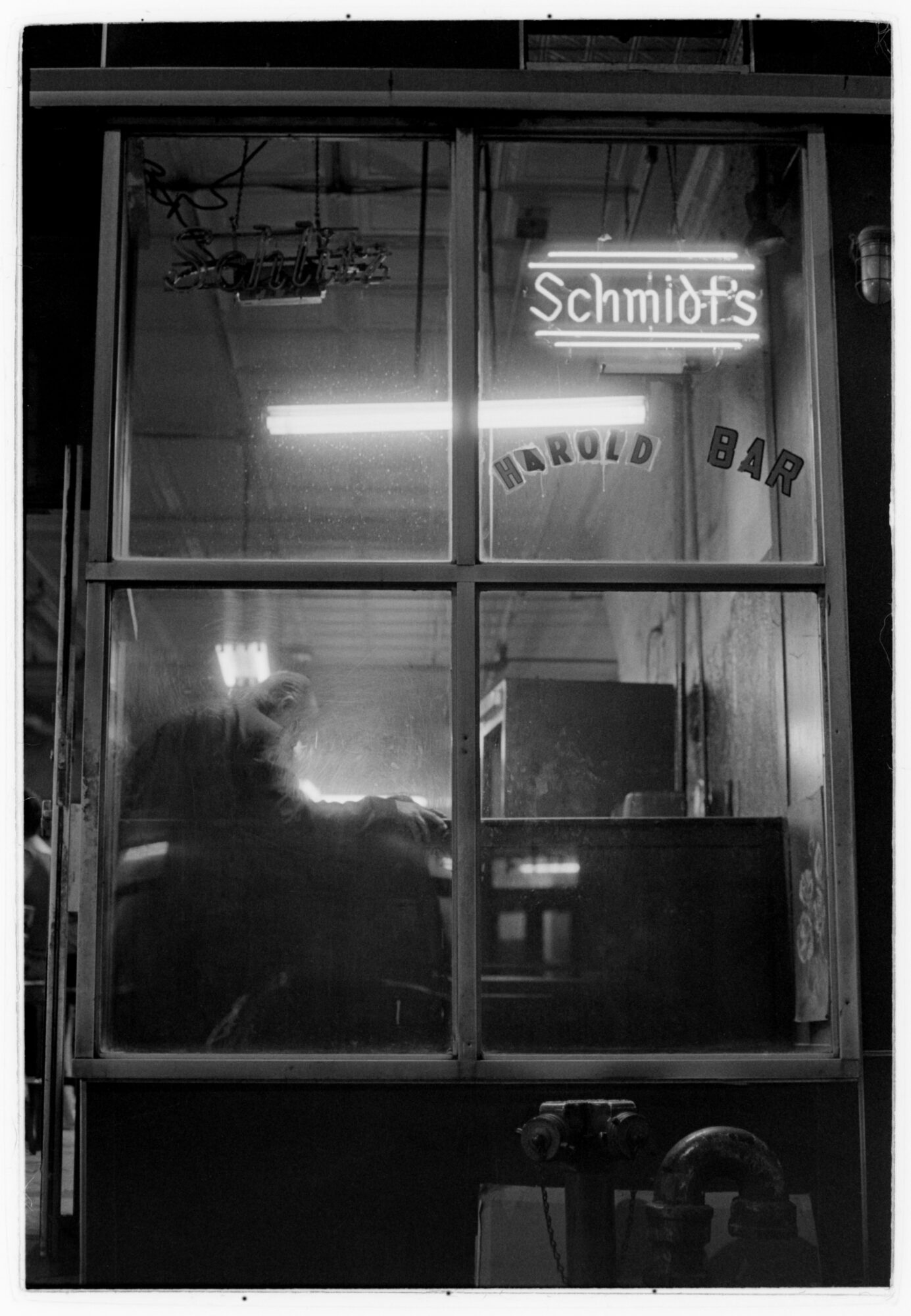 Through a bar window at night, beneath a lit Schmidt's sign and an unlit Schlitz sign, a hunched figure leans on the bar with their head resting on one hand.