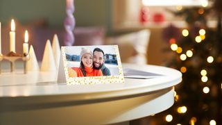 A printed Christmas card featuring a photo of a smiling couple in winter hats is displayed on a white table next to lit candles, with a blurred Christmas tree in the background.