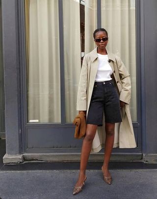 Female fashion influencer Sylvie Mus poses on a sidewalk in Paris wearing rectangular sunglasses, a collared trench coat, white T-shirt, faded black denim Bermuda shorts, a tan suede clutch, and snakeskin mule pumps