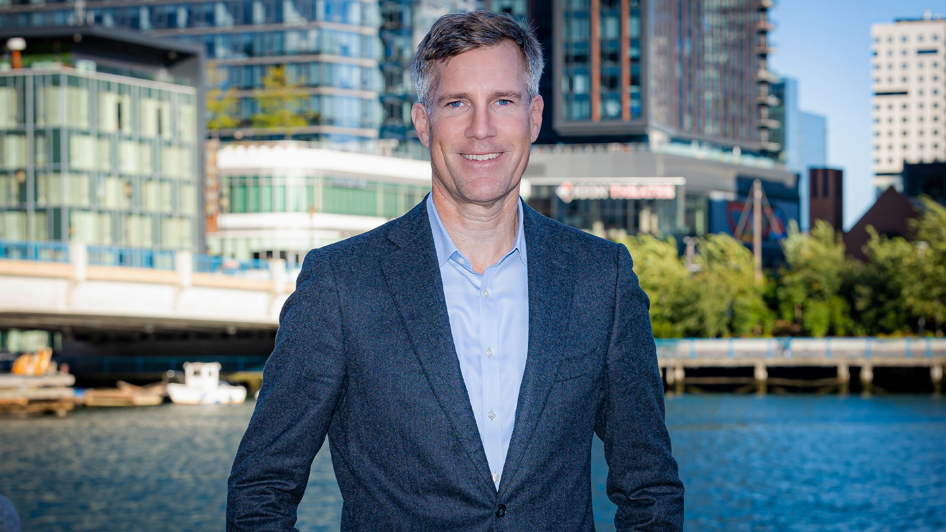 Darktrace CEO Ed Jennings wearing a navy suit and light blue shirt, with water and high rise buildings in background.