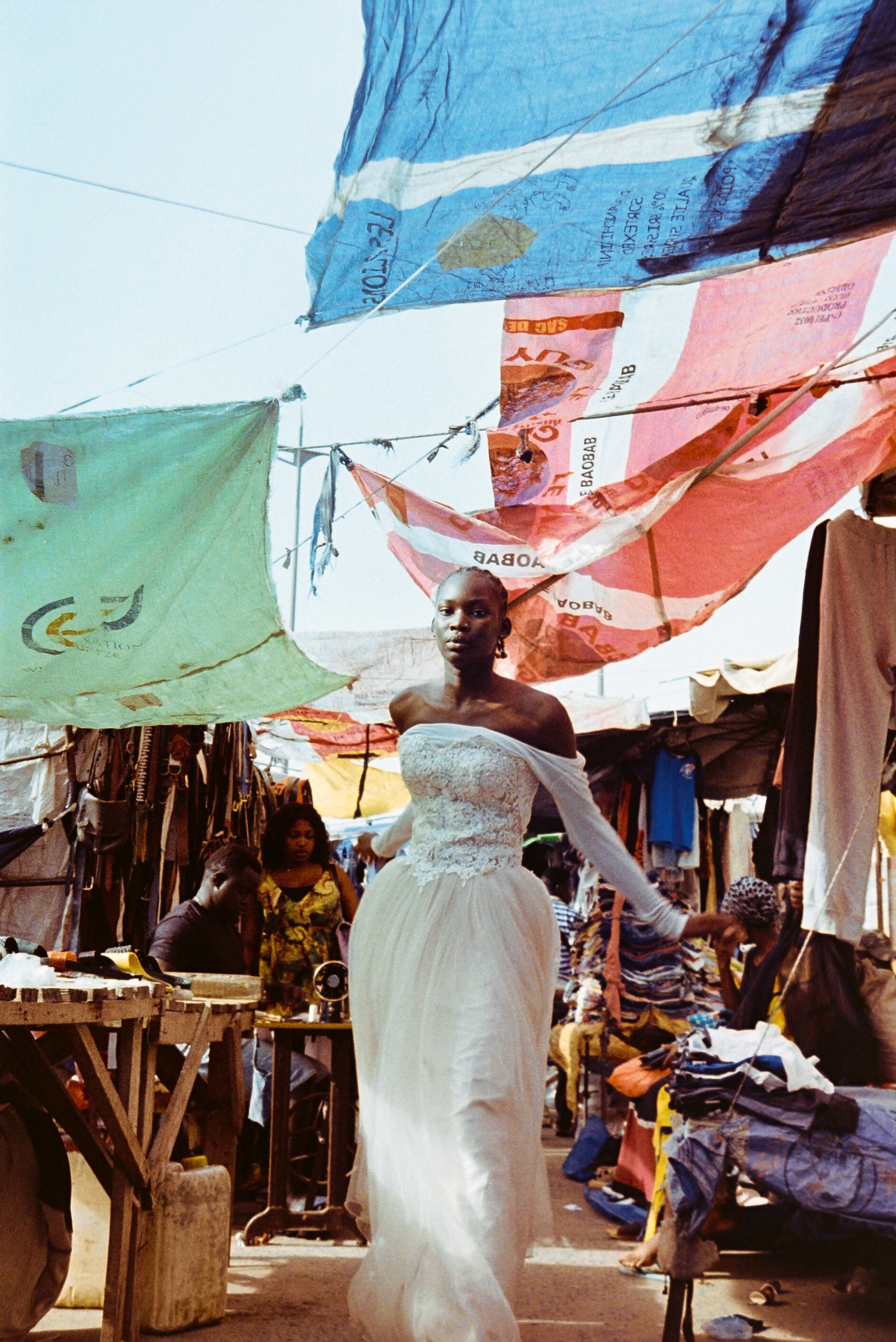 A woman in a lace and tulle wedding dress stands in a crowded market stall, framed by hanging tarpaulins and a working sewing table.