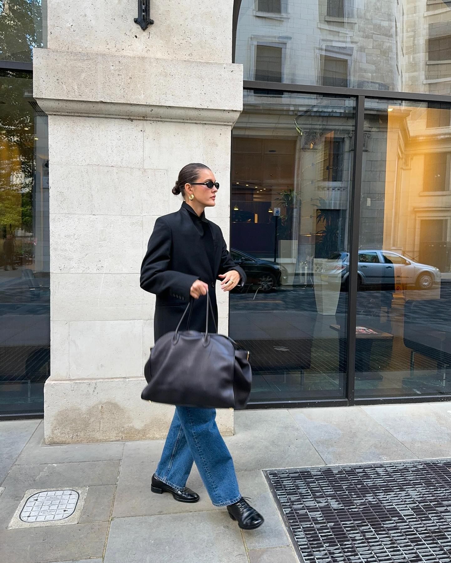 Influencer @sobalera stands outside wearing Oxford shoes, blue jeans, an oversized blazer and a large black handbag.