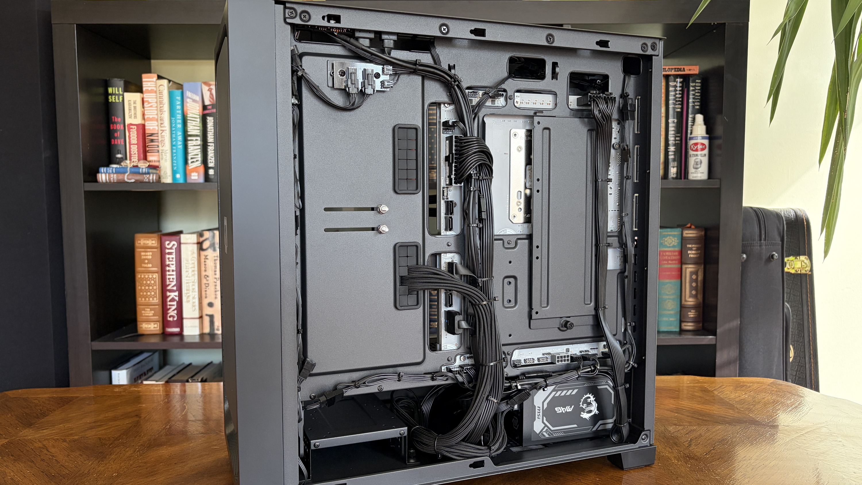 The image shows an open MG-1 MK.II computer case on a wooden table, revealing neatly organized cables and components. Bookshelves with books appear in the background.