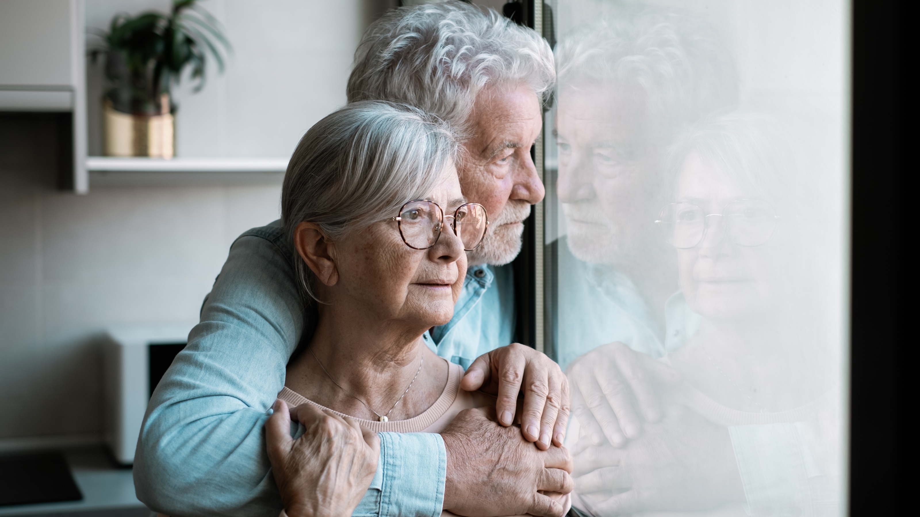 A worried older couple hold each other while looking out the window of their home.