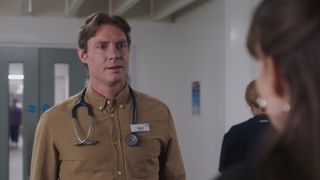 Max Brown as consultant Henry wearing a tan shirt with a stethoscope around his neck talking to Jodie.