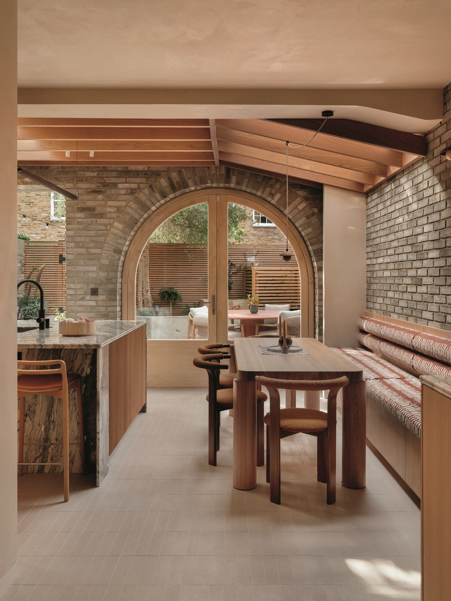 modern kitchen extension with brick walls and floors, exposed beam ceilings, banquette seat running along one wall next to a dining table, marble kitchen island, and arched doors leading to garden