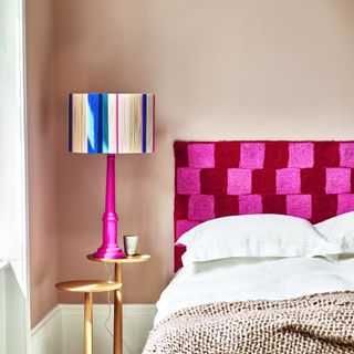 A light brown-painted bedroom with a tonal knitted throw on the bed and a contrasting striped pink table lamp and crochet headboard in pink and red