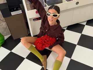 Fashion influencer Vicky Montanari wearing a suede burgundy jacket styled with a red clutch, butter yellow sunglasses, olive green socks, and brown suede boat shoes.