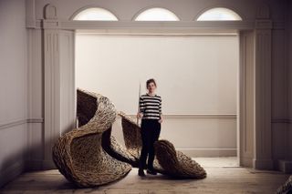 A young woman dressed in a stripy black and white t-shirt and black trousers stands next to a large-scale installation made of woven tree branches in a wooden-drenched gallery room painted white.