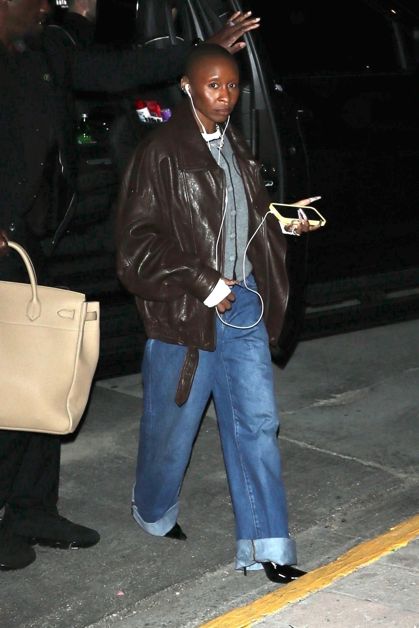 Cynthia Erivo wearing a brown leather bomber jacket, blue jeans, and a grey sweater