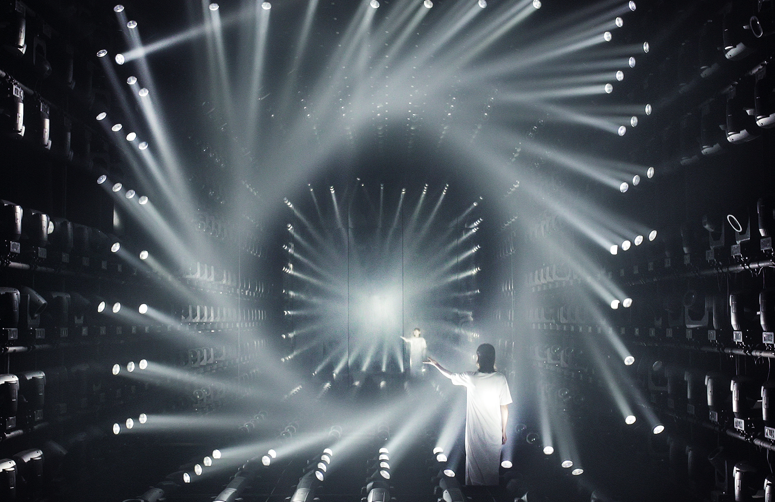 Inside a room filled with lights forming light beams and two people standing with their hands to the side.
