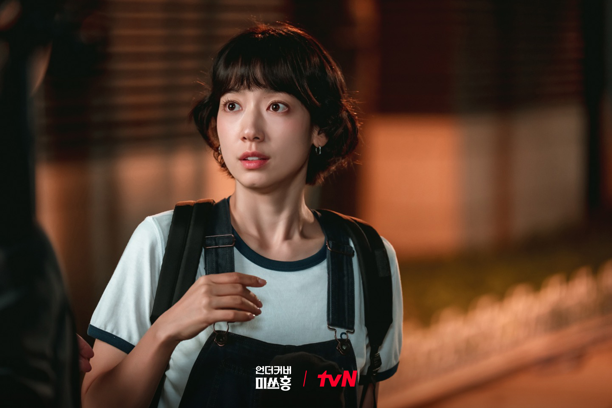 A Korean woman in her 30s (actress Park Shin-hye) wears a short, youthful hairstyle, t-shirt, overalls, and backpack as she stands with an unseen person outdoors at night. A still from the K-drama 'Undercover Miss Hong.'