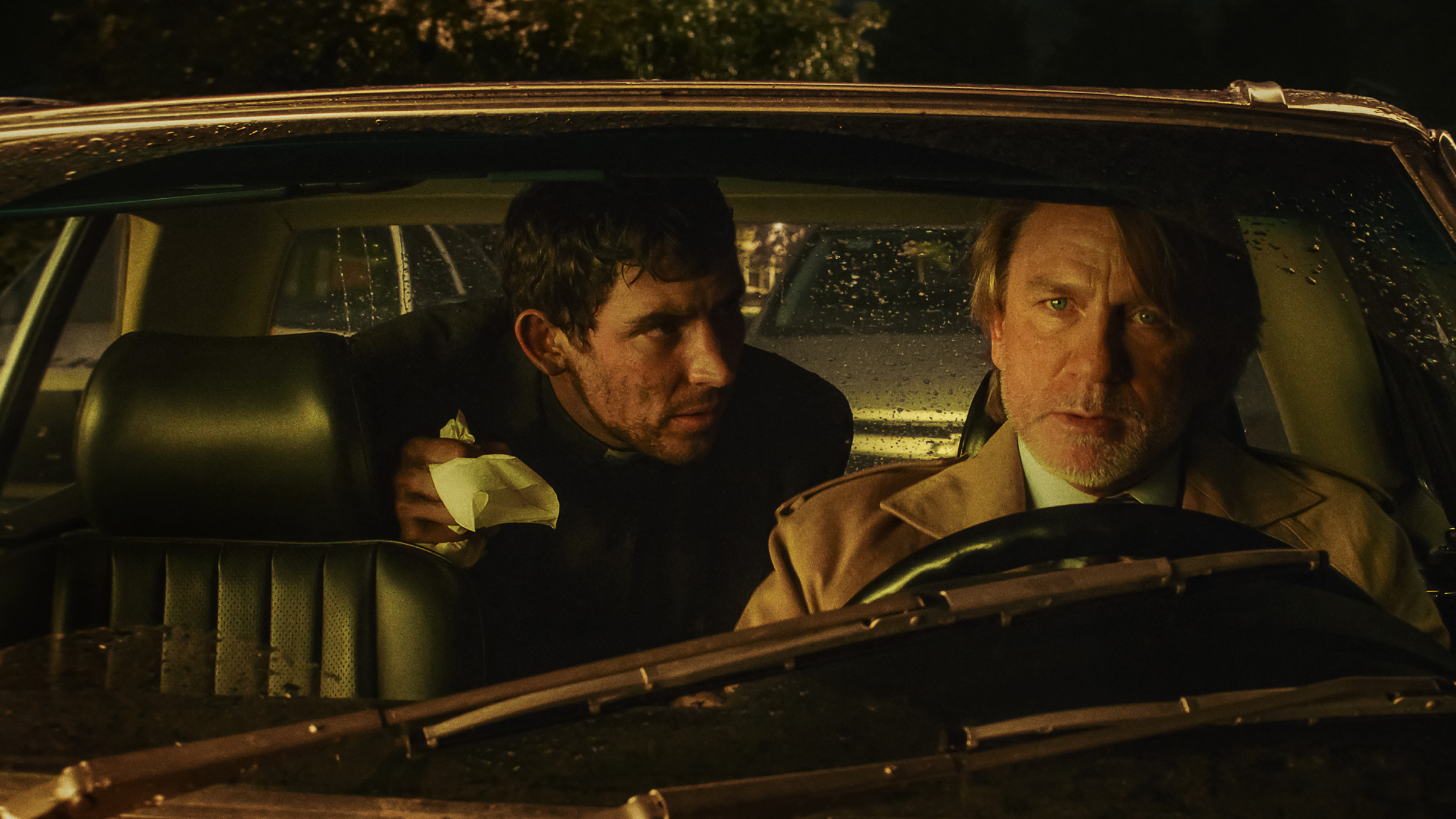josh oconnor as a priest in the back of a car talking to daniel craig as benoit blanc in the front seat in wake up dead man