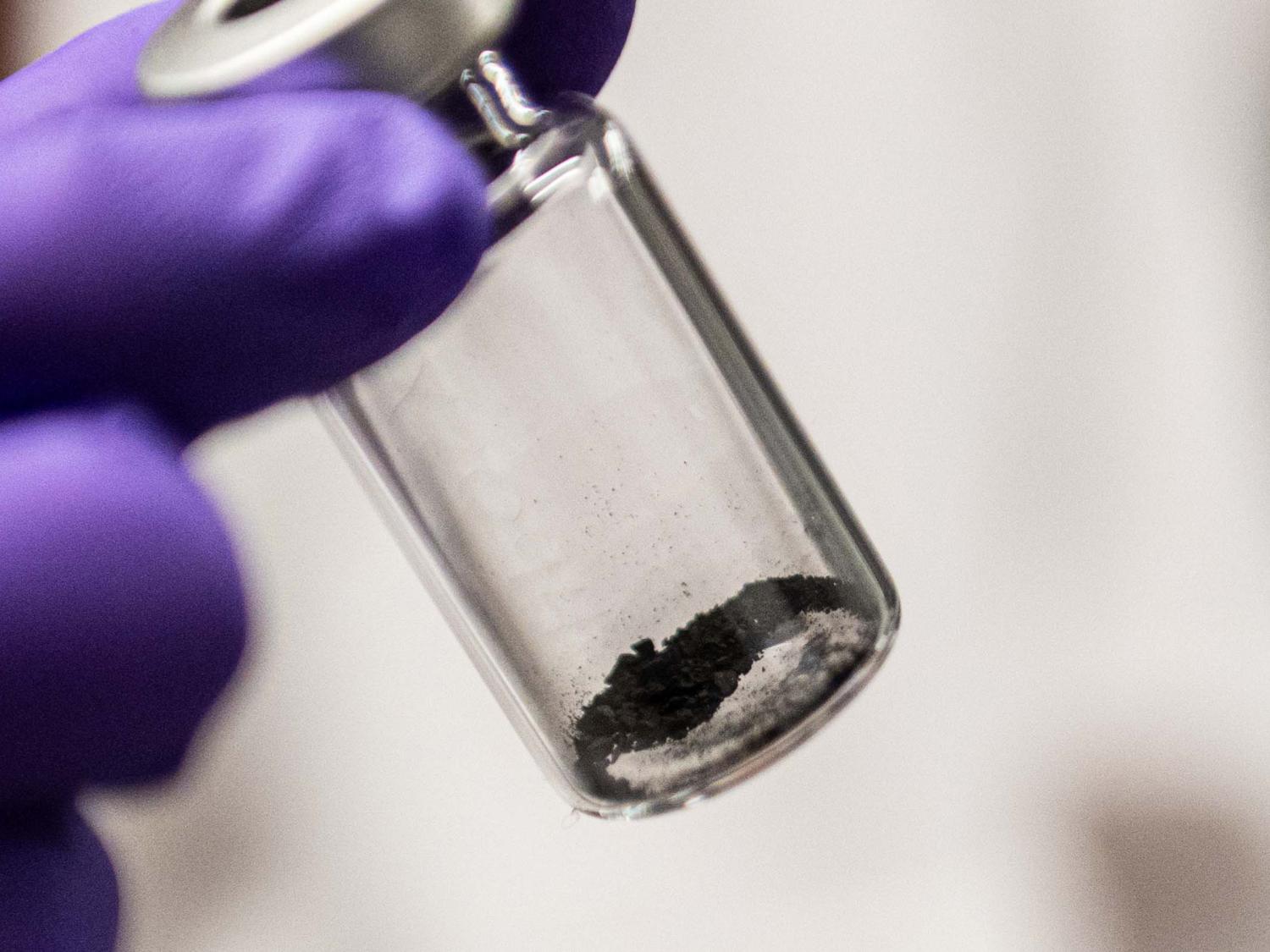 يد أرجوانية بقفاز تحمل أنبوبًا شفافًا بداخله القليل من الغبار الرمادي الداكن.