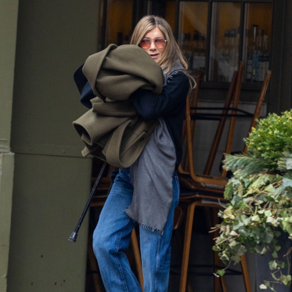 Jennifer Aniston wears puddle jeans with a navy knit and an olive green coat.