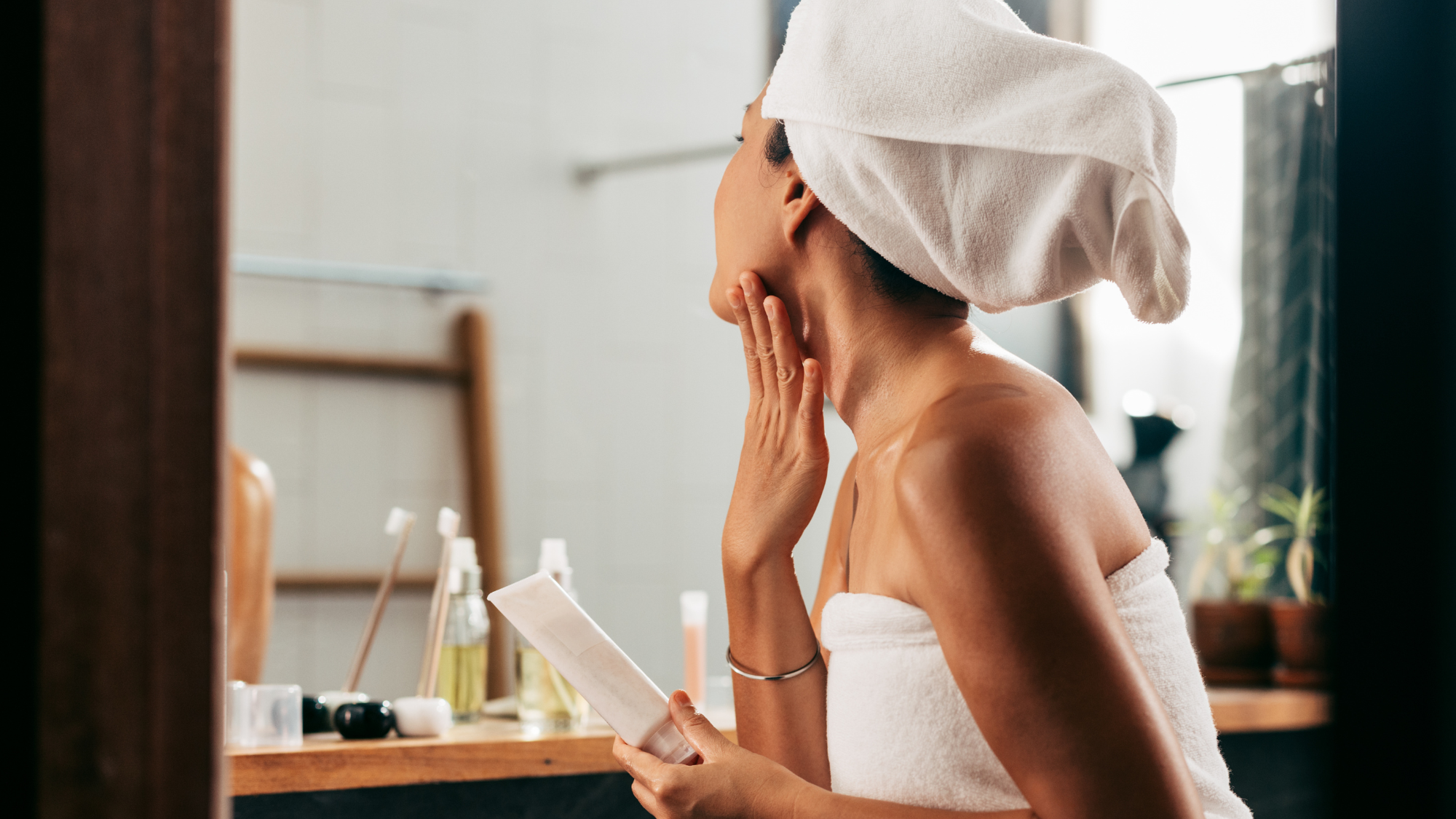 Woman Wrapped In A Towel Looking Herself In The Mirror While Applying Some Creme On Her Face After Taking A Bath