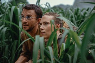 jonathan bailey wearing tiny glasses and scarlett johansson crouched low in the grass in the movie jurassic world rebirth