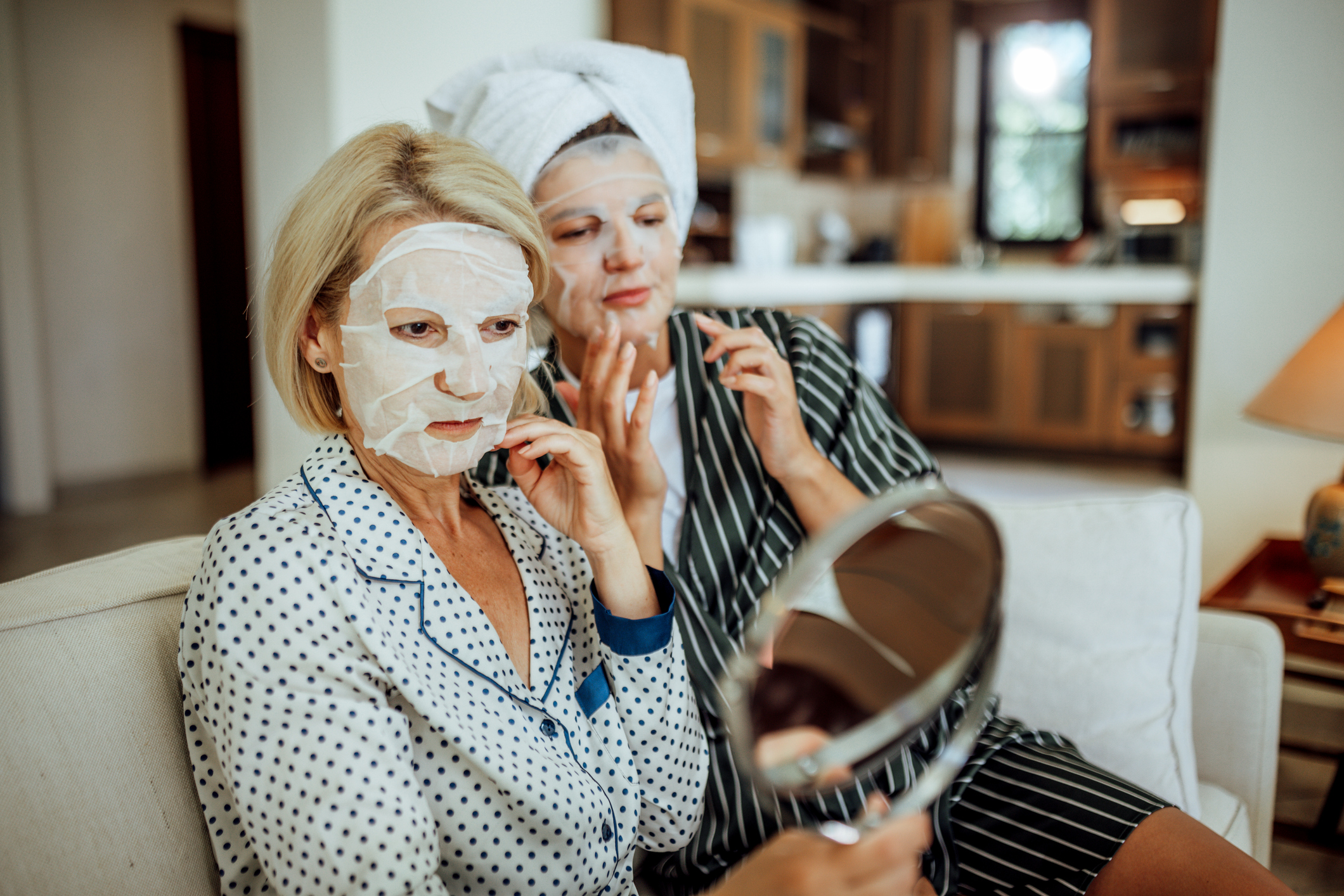 An adult daughter and mother wear sheet masks sitting on the couch.