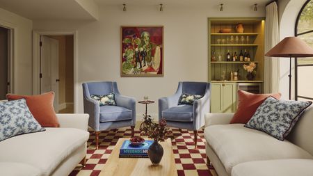 Colorful living room with two sofas and armchairs, above red-and-white checkered floor. A coffee table is pictured in the center of the room, and a large framed painting and a built-in shelving unit is pictured on the back wall.