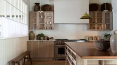 Neutral kitchen with large range cooker and wall mounted wooden cabinets with uplighting. There is a wooden island off center, to the front, and a narrow bench on the left with crittal white windows and translucent window treatments