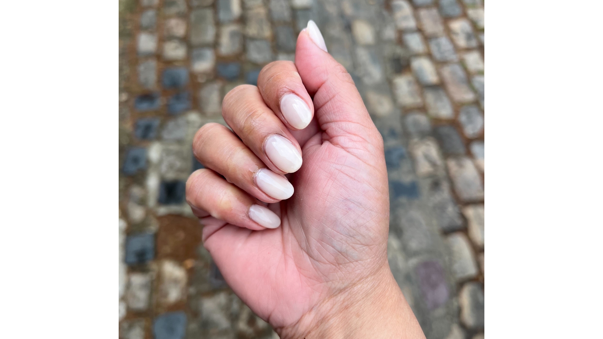 A close-up of Digital Beauty eCom Editor, Aleesha Badkar's hand seen with a creamy, coconut milk-inspired manicure/ taken above a pavement background