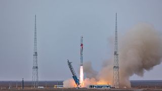 a rocket rises as fire blasts from the bottom. brown smoke rises against a grey sky.