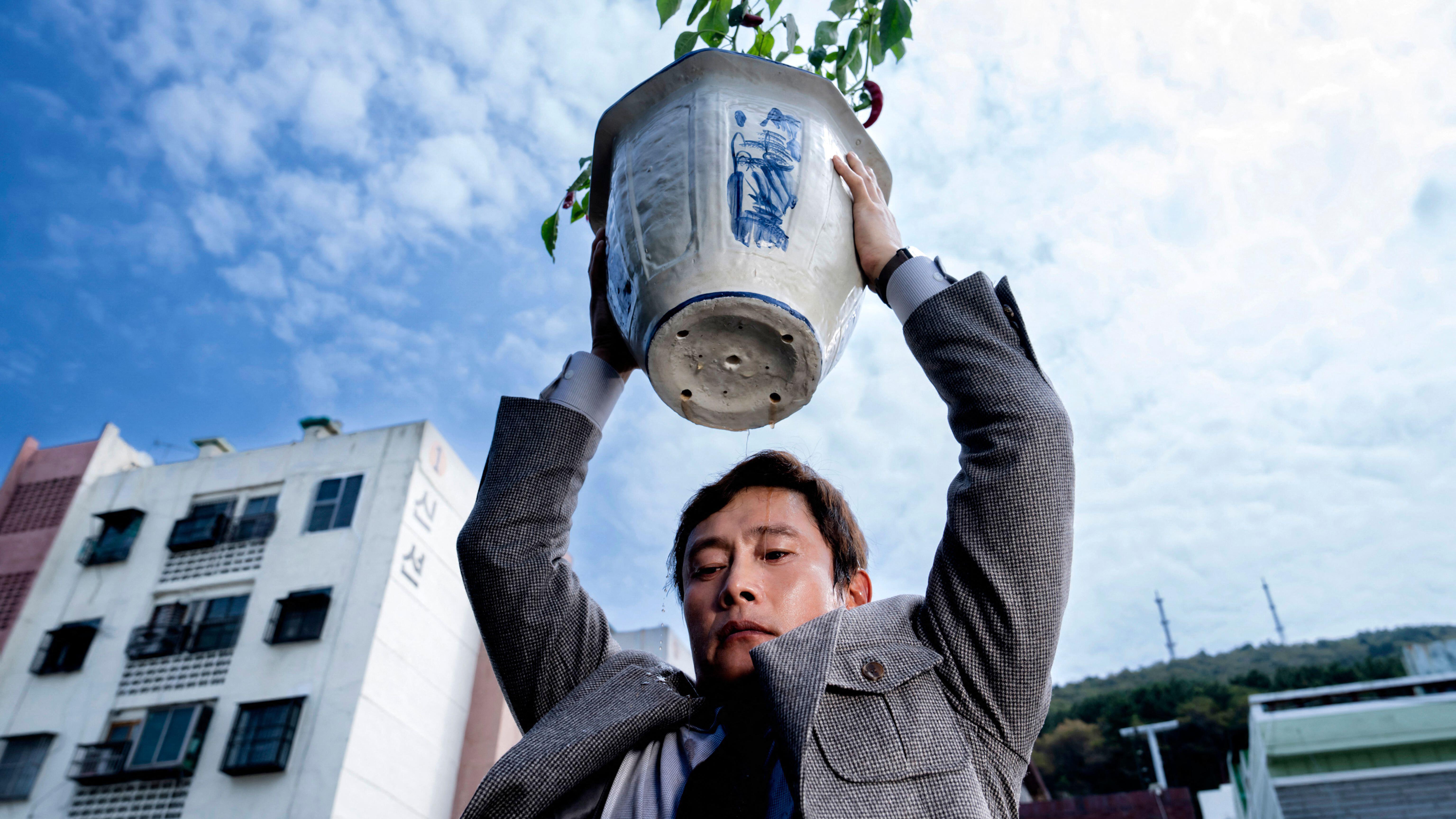 Man-su (Lee Byung-hun) holds a plant pot above his head in a scene from "No Other Choice"