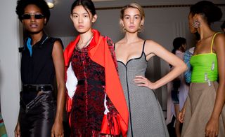 Models wear black top and leather skirt, red dress and jacket, grey dress, and green fluorescent top with beige skirt
