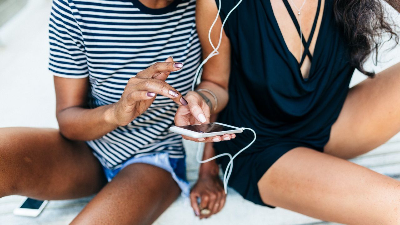 Two friends sitting on stairs listening music together with earphones and smartphone, partial view - stock photo