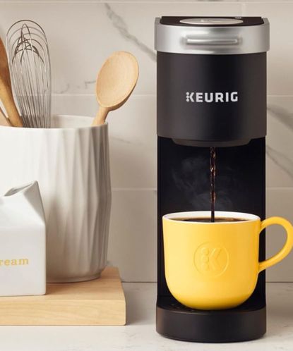 A yellow mug beneath a Keurig coffee maker; to the left, a jar of kitchen utensils and a jar of cream against a white wall. 