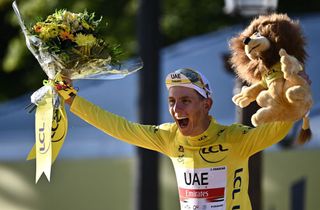 Tadej Pogačar celebrating his Tour de France win, wearing his yellow jersey and holding up a large bouquet of flowers and a stuffed lion