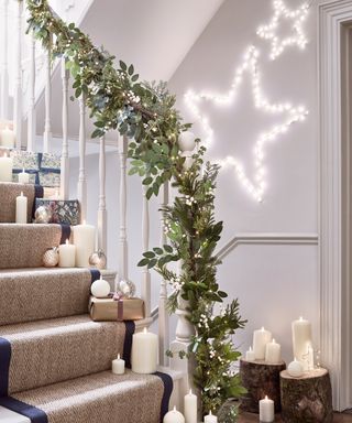 a festive staircase with faux candles, presents and baubles on top of the rattan and ribbon stair runner, a garland down the banister, and star lights on the white walls