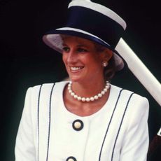 Princess Diana wears a white blazer with navy blue piping and buttons with a matching hat as she sits with a young Prince Harry in a suit