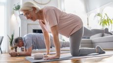 older woman and man performing core exercises in a living room setting. she is in the foreground on her hands and knees with one leg extended back and he's in the background in a plank position.