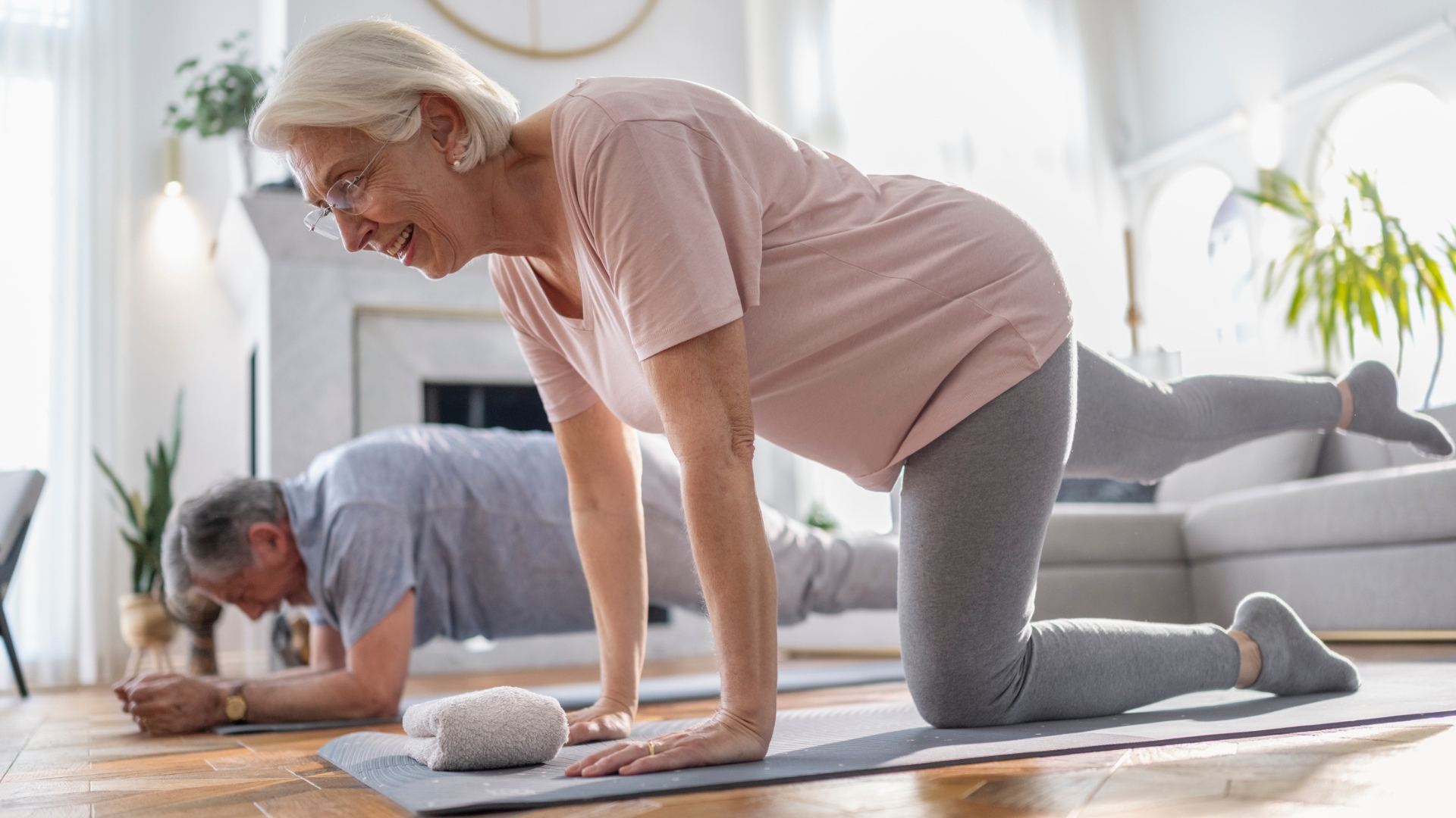 older woman and man performing core exercises in a living room setting. she is in the foreground on her hands and knees with one leg extended back and he&#039;s in the background in a plank position. 