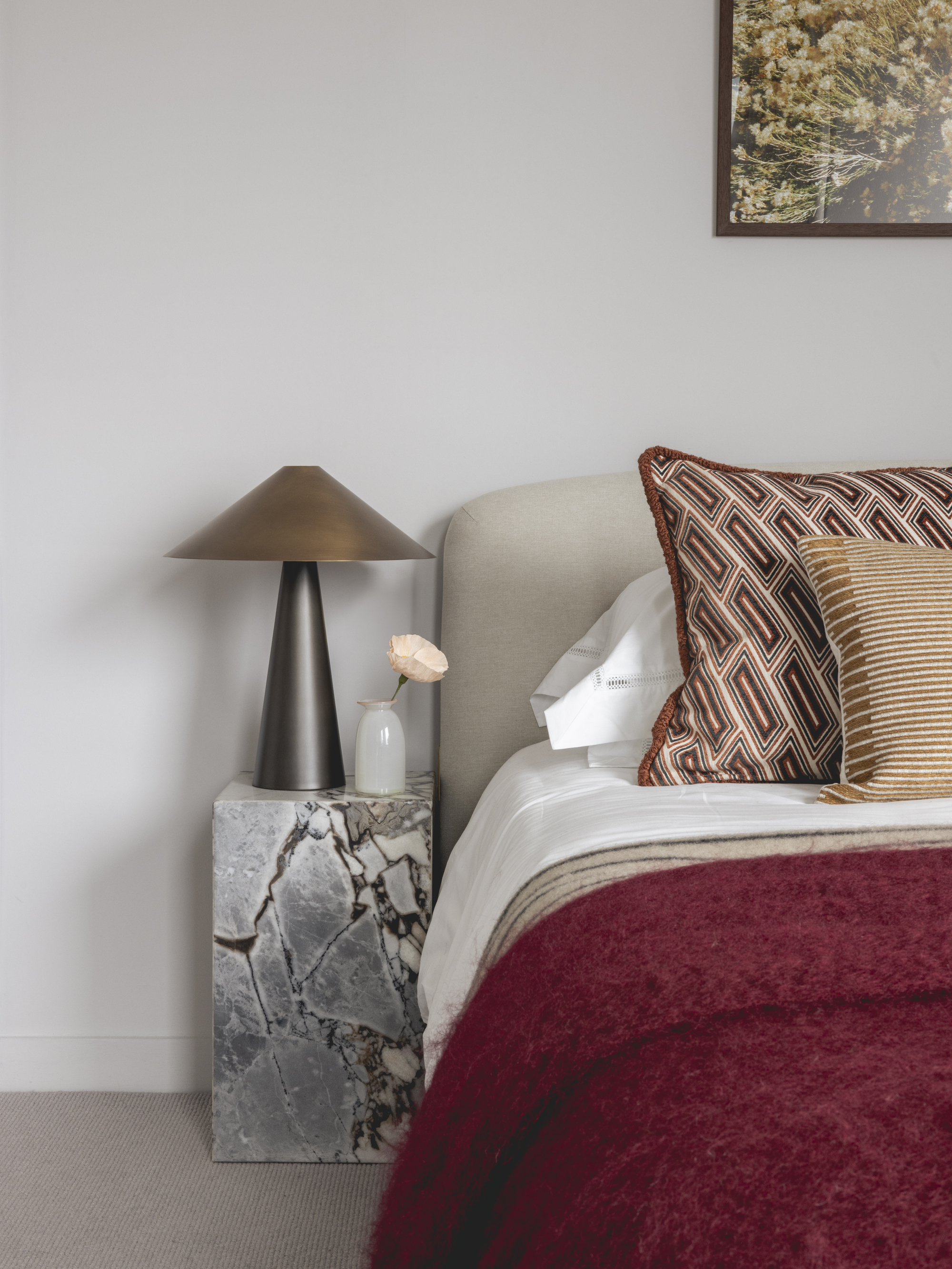closer up of bedroom with white walls, artwork hung above bed, beige upholstered headboard, white pillowcases and sheets, red blanket, red patterned cushion and striped cushion, marble plinth side table, with bronze lamp and vase holding a flower stem