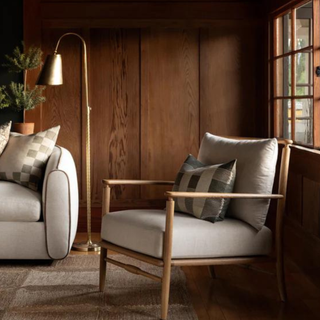 A gray and wood lounge chair with a throw pillow, next to a window and a gold standing lamp.