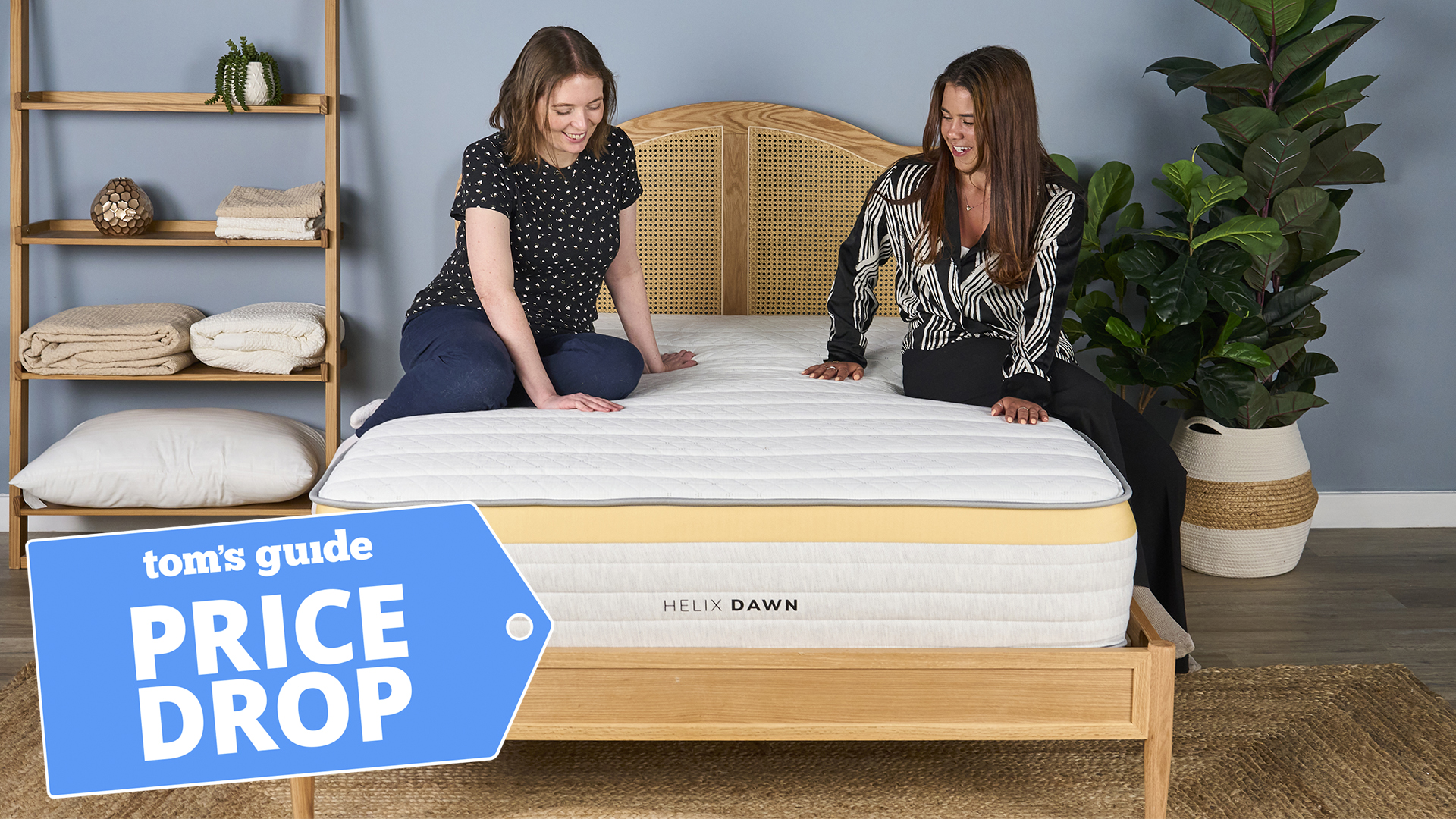 Two women sit on the Helix Dawn mattress on a bed frame in a bedroom, testing the comfort and feel of the mattress. A Tom's Guide Price Drop deals badge in the bottom left corner
