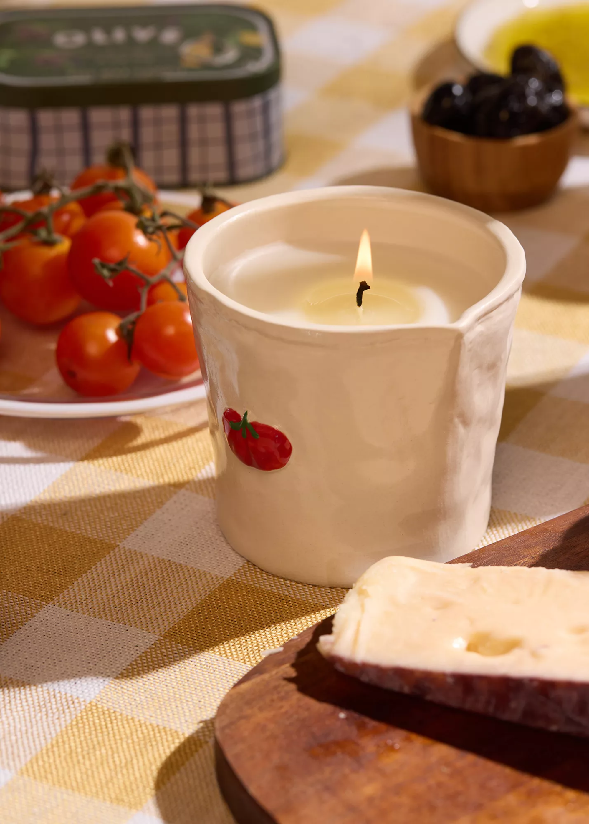 A tomato-scented ceramic candle on a checkerboard tablecloth by a plate of vine tomatoes and a board of cheese