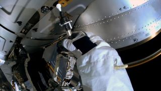 A first-person view from a helmet-mounted camera showing an astronaut's gloved hand holding a swab outside of a space station.