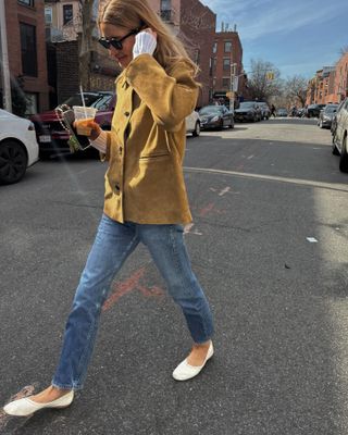 Candid photo of a NY woman in a tan suede jacket, straight jeans and white ballet flats drinking an iced coffee