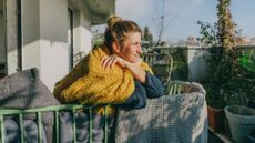 A woman leans over a balcony edge with a blanket across her shoulders, squinting into the distance. Behind her we see plants and a city skyline. 