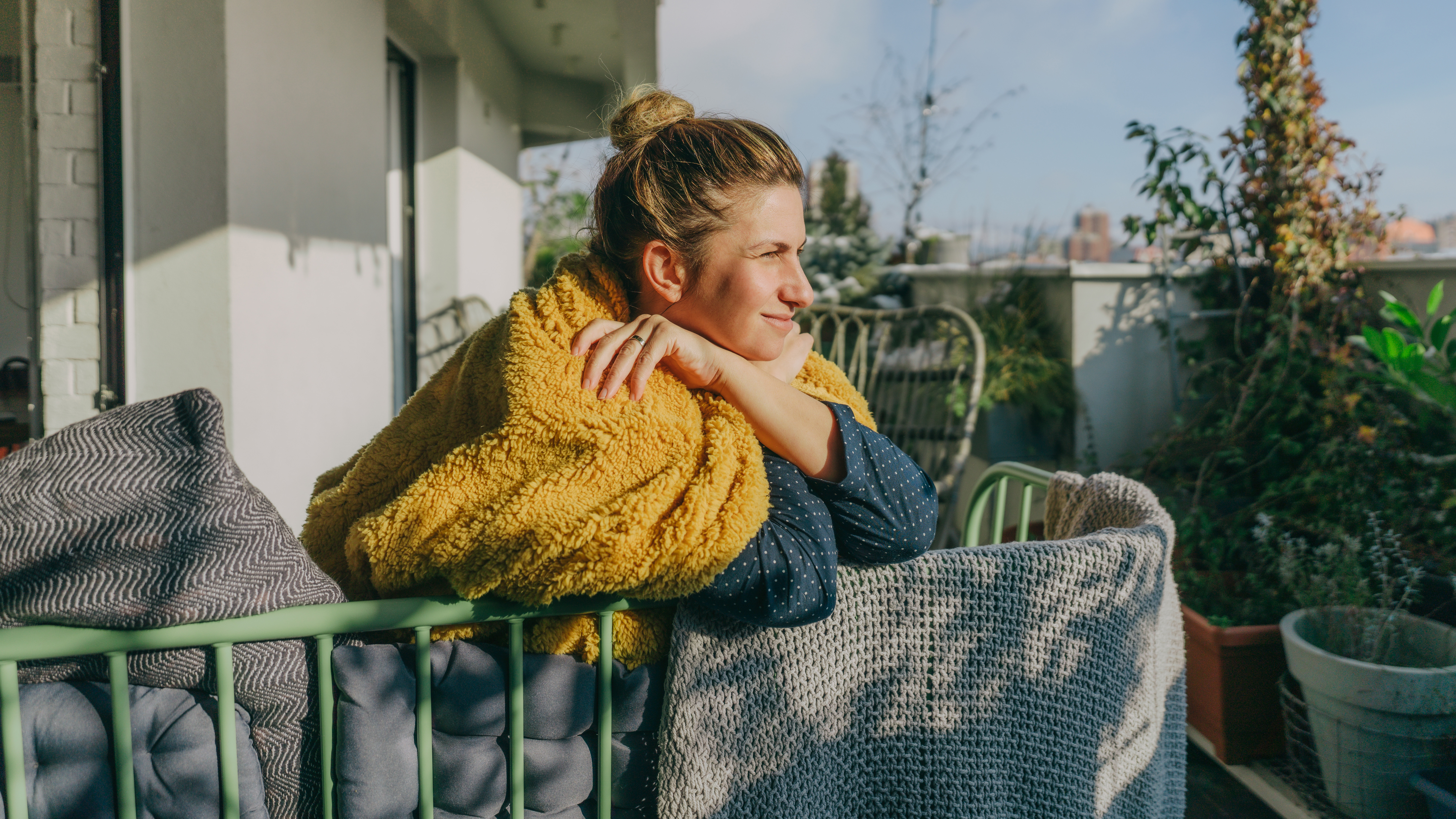 A woman leans over a balcony edge with a blanket across her shoulders, squinting into the distance. Behind her we see plants and a city skyline. 