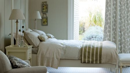 Neutral bedroom with patterned wallpaper, white four-poster bed, and large window with garden seen beyond and patterned curtains.