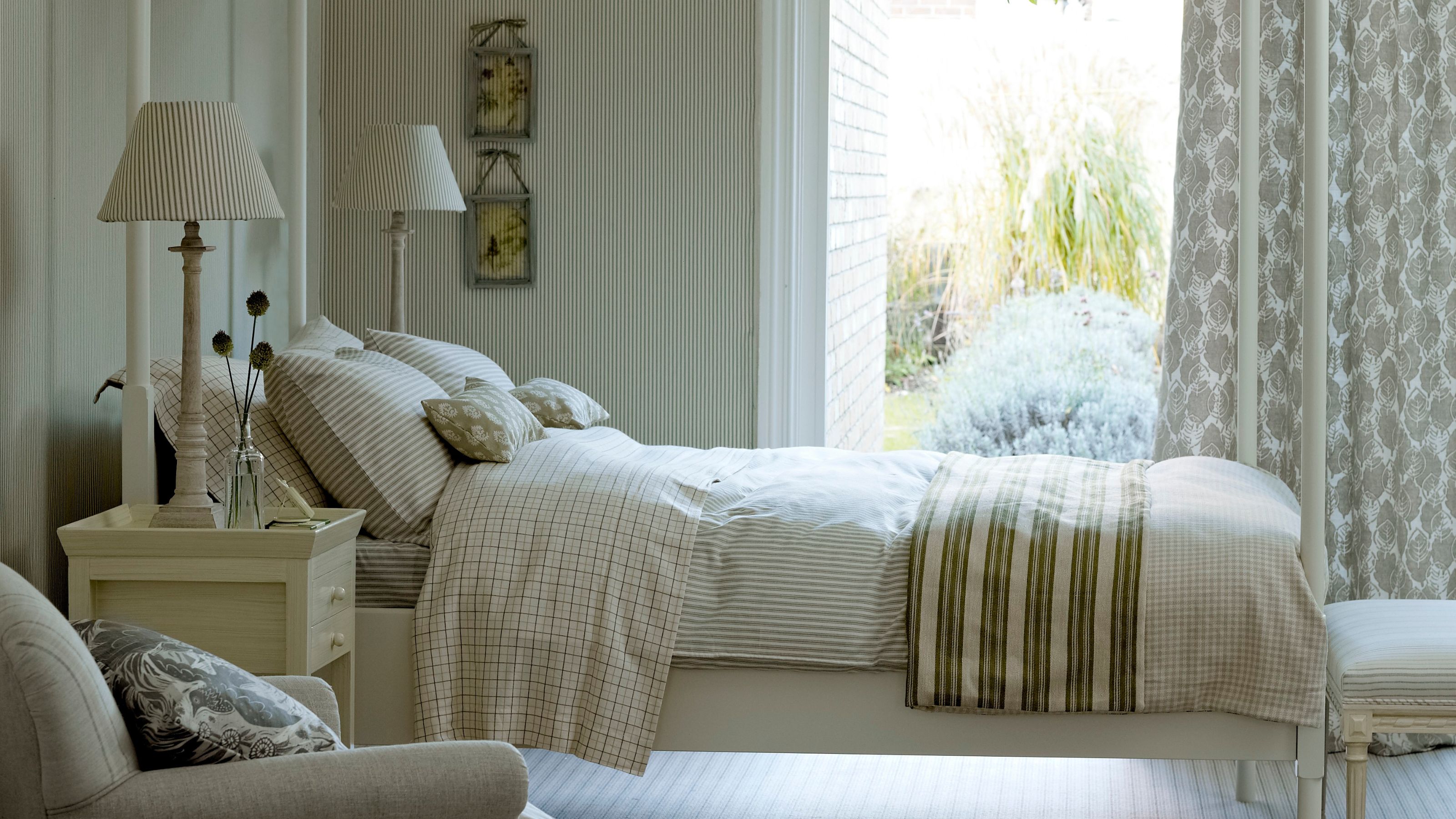 Neutral bedroom with patterned wallpaper, white four-poster bed, and large window with garden seen beyond and patterned curtains.