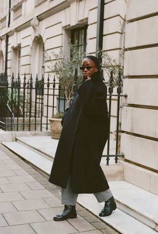 A woman wearing a black headband, black sunglasses, and a black funnel neck coat with gray knit pants and black riding boots.