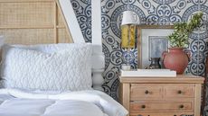 A Coop Sleep Goods pillow on a bed against a wooden frame; to the right, a nightstand, topped with a lamp, a vase, and books, against blue and white patterned wallpaper.